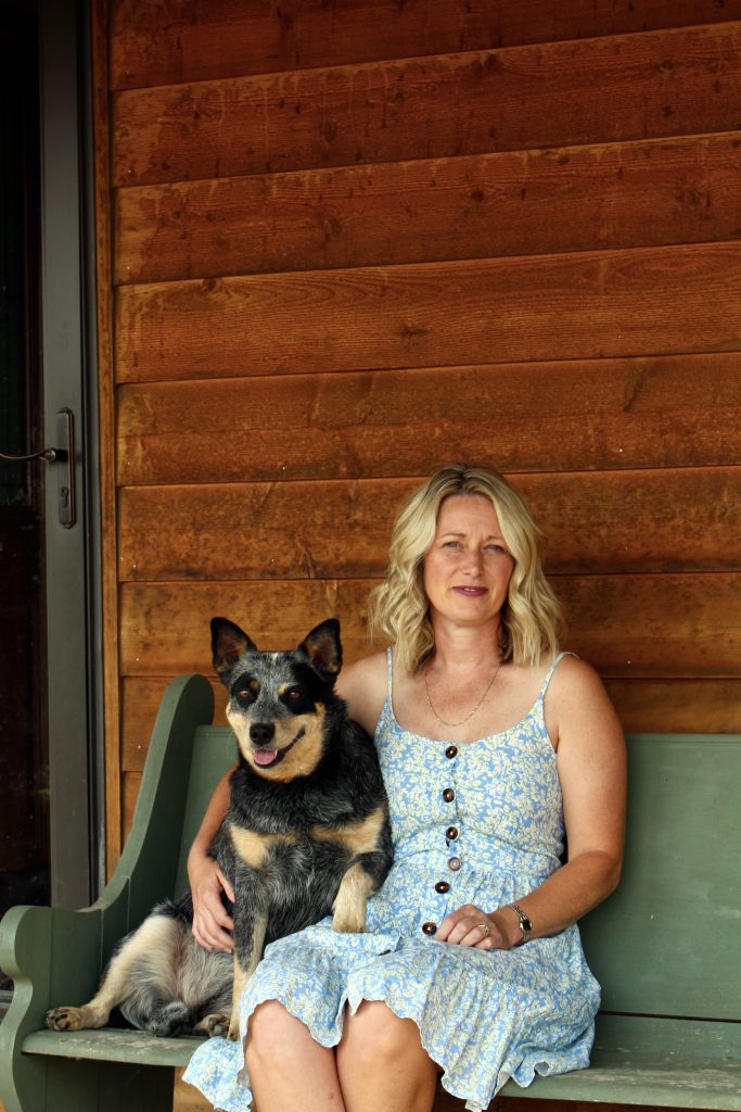 Woman sitting in sundress on green bench with arm around cattle dog.  About Season